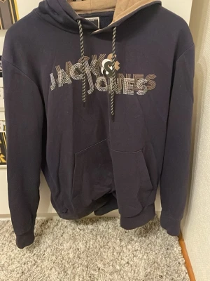 Mörkblå hoodie från Jack & Jones - Snygg mörkblå hoodie från Jack & Jones med stor logga framtill i vitt och beige. Hoodien har en kontrastfärgad beige insida i huvan, dragsnören och en stor magficka. Perfekt för en avslappnad och trendig stil.