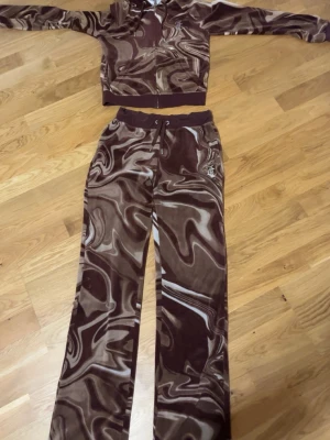 Brunt mönstrat Juicy Couture set - Säljer ett brunt loungewear set från Juicy Couture med swirl-mönster. Setet består av en zip-hoodie med huva och matchande byxor med dragsko i midjan. Både hoodie och byxor har Juicy-loggan i strass på ryggen och bakfickan. Mjukt velourmaterial för maximal komfort.