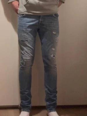 Blå Dondup jeans med slitningar - Säljer ett par blå Dondup jeans med slitna detaljer och hål på benen. Jeansen har en tajt passform och klassisk femficksdesign. Perfekta för dig som gillar en trendig och avslappnad stil.