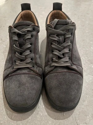 Mörkgrå Louboutin  - Snygga mörkgrå sneakers i mocka med klassisk snörning och rund tå. Skorna har en unik röd sula som sticker ut och ger en cool kontrast till den matta ovandelen. Perfekta för dig som gillar stilrena men ändå unika detaljer. Släppt lite i sömnen, se bilder, men inget som syns när man har dem på sig. Därav lägre pris.