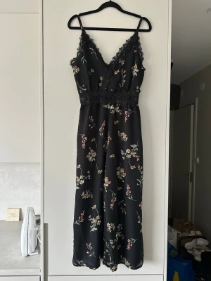 Svart blommig långklänning med spets - Svart långklänning med tunna axelband, blommigt mönster och v-ringad spetskantad urringning. Klänningen har spetsdetalj i midjan och ett flowy fall som ger en romantisk vibe. Perfekt för dig som gillar feminina detaljer och stilren design.