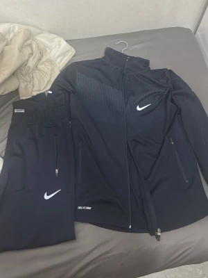 Nike svart tracksuit i polyester M - Nike tracksuit i svart med jacka och byxor i 100% polyester. Jackan har dragkedja, hög krage och vita Nike-loggor på bröstet och byxbenet. Byxorna har resår i midjan och dragkedjor vid fickorna. Perfekt för träning eller chill.