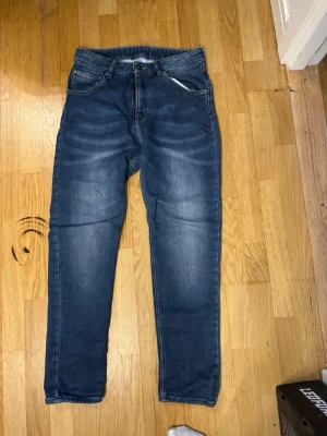 Blå jeans med raka ben för barn  - Säljer ett par klassiska blå jeans med raka ben och normal passform. Jeansen har fem fickor, knappgylf och snygga slitningar framtill för en avslappnad look. Materialet är tåligt denim och färgen är mellanblå med ljusare partier.