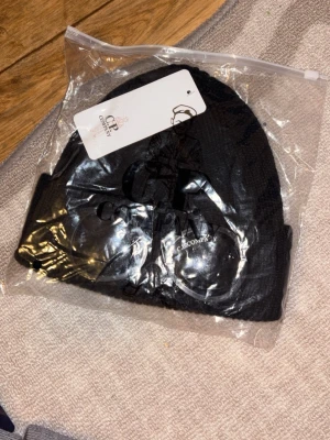 Svart mössa från C.P. Company - Svart ribbstickad mössa från C.P. Company med ikoniska inbyggda glasögon framtill. Klassisk uppvikt kant och stilren design, perfekt för dig som gillar streetwear och vill sticka ut med en unik accessoar.