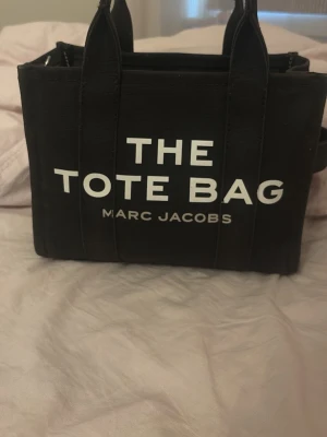 Svart tote bag från Marc Jacobs - Säljer en svart tote bag från Marc Jacobs med tydlig vit text på framsidan. Väskan har två rejäla handtag och är tillverkad i slitstark bomull. Perfekt storlek för att få plats med allt du behöver och har en cool, stilren design.