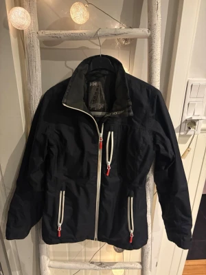 Svart vindjacka från Helly Hansen - Svart vindjacka från Helly Hansen med vita dragkedjor och röda detaljer. Jackan har hög krage, två fickor framtill och HH-logga på ryggen. Tillverkad i slitstark polyester och perfekt för blåsiga dagar.