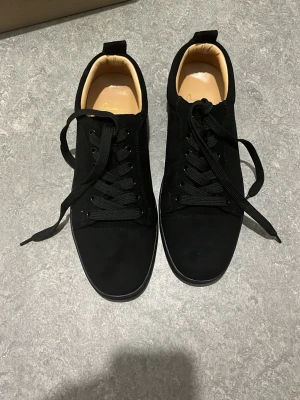 Svarta sneakers från Christian Louboutin - Stilrena svarta sneakers från Christian Louboutin med klassisk röd sula och svarta skosnören. Ovandelen är i svart material med diskret design och insidan är ljusbeige. Perfekta för dig som gillar exklusiva och trendiga skor.