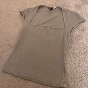 Beige topp med fyrkantig ringning H&M - Snygg grön topp från H&M med korta ärmar och unik fyrkantig ringning. Tillverkad i mjuk bomullsmix som sitter skönt mot kroppen. Perfekt att styla med jeans eller kjol för en clean look.