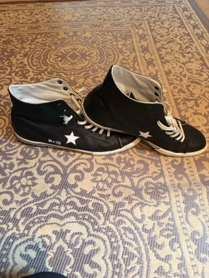 Höga converse - Säljer ett par svarta high-top sneakers med vita stjärnor på sidan och vita snören. Skorna har vit sula och insida, samt en klassisk rund tå. Riktigt feta skor som passar med allt, passar 41-42. Hör av er vid minsta fundering!
