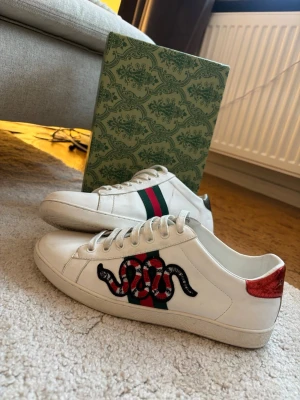 Gucci Ace sneakers med orm - Gucci Ace sneakers i vitt skinn med ikonisk orm-brodyr på sidan, gröna och röda ränder samt röd metallic-detalj på hälen. Klassisk låg modell med snörning och rund tå. Kommer med originalförpackning och tillbehör.