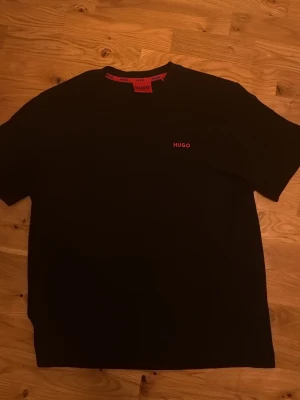 Svart t-shirt från HUGO - Snygg svart t-shirt från HUGO med röd logga på bröstet och röd etikett i nacken. Helt ny, använd 1-2 gånger. Säljer då den var för stor.