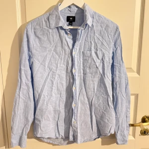Ljusblå skjorta från H&M - Klassisk ljusblå skjorta från H&M i regular fit. Skjortan har lång ärm, bröstficka och knappar framtill. Tillverkad i mjuk bomull som känns skön mot huden. Perfekt till jeans eller chinos för en clean och enkel look.