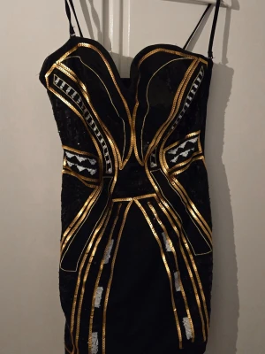 H&M Svart och guld kort klänning - Snygg svart kort klänning drån H&M med tunna axelband och detaljer i guld och silver. Klänningen har ett geometriskt mönster med paljetter som ger en glittrig effekt. Perfekt för dig som vill sticka ut med en unik och trendig look. Nypris 595 kr