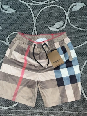 Burberry badbyxor med rutigt mönster - Snygga badbyxor från Burberry med klassiskt beige, svart, vit och röd rutigt mönster. De har elastisk midja med dragsko och sidofickor samt en bakficka. Tillverkade i ett lätt och snabbtorkande material, perfekta för stranden eller poolen.