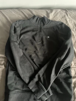 Svart half-zip tröja från Calvin Klein - Svart sweatshirt med half-zip och hög krage från Calvin Klein. Tröjan har diskret CK-logga broderad på bröstet och ribbade muddar vid ärmslut och nederkant. Perfekt för en clean och stilren look. Storlek170-180cm