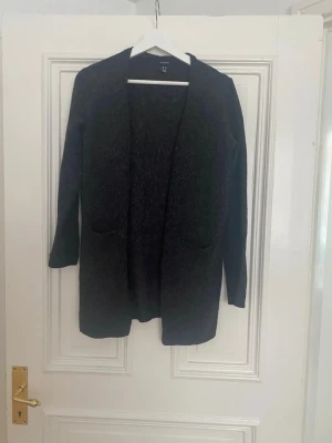 Mörkgrå lång kofta från Lindex - Mysig mörkgrå lång kofta från Lindex med öppen front och två fickor framtill. Perfekt att slänga över en t-shirt eller topp för en avslappnad look. Koftan har långa ärmar och en rak passform.