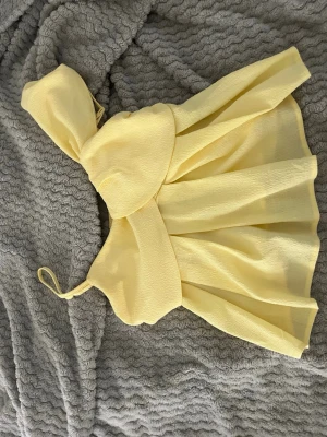 Gul croppad topp med rosettdetalj - Söt gul croppad topp med stor rosettdetalj framtill och breda axelband. Toppen har en luftig och utsvängd passform, perfekt för varma dagar. Tillverkad i ett lätt och strukturerat tyg som ger en somrig känsla.