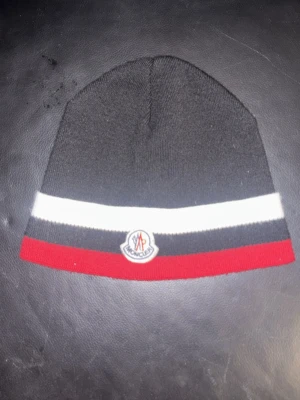 Svart Moncler mössa - Snygg svart mössa från Moncler med vit och svart rand samt röd kant längst ner. Klassisk Moncler-logga framtill. Perfekt accessoar för kalla dagar och passar till de flesta outfits. Materialet känns mjukt och bekvämt.