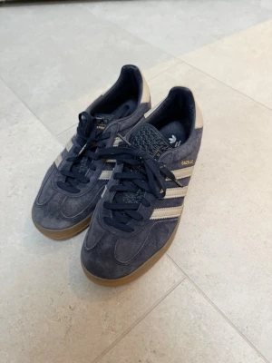Adidas Gazelle mörkblå mockasneakers - Snygga Adidas Gazelle sneakers i mörkblå mocka med klassiska vita ränder på sidorna och gummisula. Skorna har snörning och gulddetaljer vid loggan. Perfekta för dig som gillar retrostil och vill ha något bekvämt och stilrent.