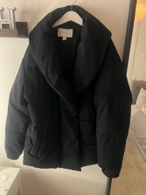 Svart pufferjacka från Vila - Snygg svart pufferjacka från Vila med oversized krage och quiltad design. Jackan har en loose passform och är perfekt för kyliga dagar. Tillverkad i polyester för en mjuk och varm känsla. Enkel att matcha med olika outfits.