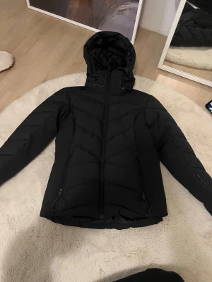 Svart skidjacka - Snygg svart skidjacka, knappt använd, fem fickor och luvan går att ta av, även knäppe runt midjan, köpt på intersport för något år sen 💕