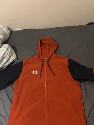 Röd och svart vindjacka från Under Armour - Snygg röd och svart vindjacka från Under Armour med huva och dragkedja framtill. Jackan har svarta ärmar med röda detaljer och en vit logga på bröstet. Perfekt för blåsiga dagar och sportiga outfits.