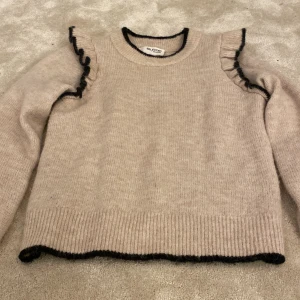 Beige stickad tröja med volang Selected Femme - Superfin beige stickad tröja från Selected Femme med svarta kontrastkanter och volangdetaljer på axlarna. Köpt för 1000kr och nästan helt oanvänd. Tröjan har rund halsringning, långa ärmar och ribbade muddar. Perfekt för dig som gillar en söt och trendig look.