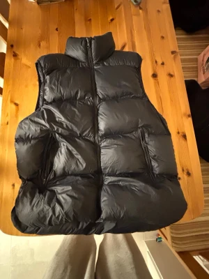 Mörkblå  dunväst från Zara - Säljer en svart, puffig dunväst från Zara med hög krage och dragkedja framtill. Västen har två praktiska fickor med dragkedja och en clean, stilren look som passar till många outfits. Perfekt för lager-på-lager under kyliga dagar.