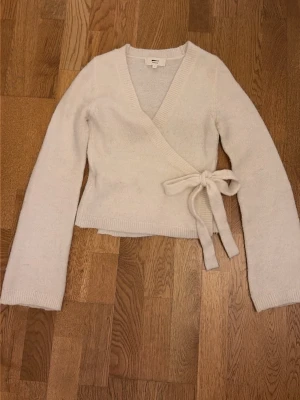 Vit omlottkofta från Gina Tricot -  vit omlottkofta från Gina Tricot i ullblandning. Koftan har långa ärmar, ribbade muddar och knyts i sidan med en bred rosett. 