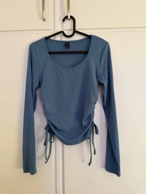 Blå ribbad topp med snörning - Säljer en blå långärmad topp från New Look med ribbad struktur och snörning i sidorna. Toppen har rund halsringning och är i ett mjukt, stretchigt material som sitter snyggt på kroppen. Perfekt att styla med jeans eller kjol.