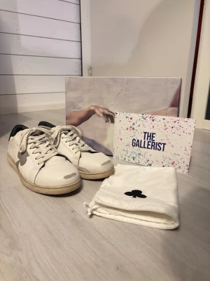 The Gallerist Vita Sneakers/Skor - Säljer ett par vita sneakers från The Gallerist med svart häl och grå detalj i form av en klöver på sidan. Skorna har snörning och platt sula. Tillverkade i skinn och har en klassisk, rund tå. Perfekta för dig som gillar stilrena och unika detaljer. Storlek 44. Skulle säga det är bra skick men tyvärr har dom spruckit lite men det är  limmat samt det är inget som syns eller stört mig (se sista bild). Allt original finns kvar så som dustbag och skokartong osv. BVSA, även byten är intressant! 🙌