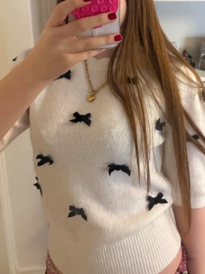 Vit stickad tröja med svarta rosetter - Supersöt vit stickad tröja med svarta rosetter över hela. Tröjan har trekvartsärmar och ribbad kant nedtill. Aldrig använd förutom för dessa bilder, köptes för 400kr.