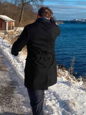 Svart kappa från J.Lindeberg - Stilren svart kappa från J.Lindeberg med klassisk krage och knappar framtill. Kappan har en rak passform och dekorativ knappdetalj i midjan. Perfekt för både kyliga dagar och som en vår jacka