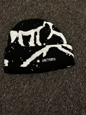 Svartvit mössa från Arc'teryx - Snygg svart mössa från Arc'teryx med vit grafisk design och uppvikt kant. Loggan är broderad framtill. Perfekt för dig som gillar streetwear och vill ha något som sticker ut. Materialet känns mjukt och stretchigt, troligen akryl eller ullmix.