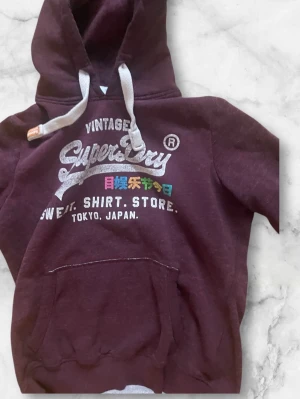 Vinröd hoodie från Superdry - Säljer en vinröd hoodie från Superdry med stor logga och text i vitt och färgglada detaljer på bröstet. Hoodien har vit dragsko, stor känguruficka framtill och mjukt material som känns skönt mot huden. Perfekt för dig som gillar streetwear och japansk-inspirerad design.