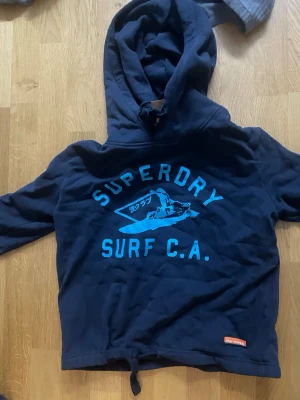 Mörkblå Superdry hoodie med tryck - Snygg mörkblå hoodie från Superdry med stort blått surf-inspirerat tryck på bröstet och justerbar dragsko nedtill. Klassisk huva och avslappnad passform, perfekt för en chill och sportig stil.
