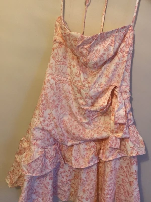 Blommig omlottklänning med volanger - Söt kortklänning med rosa och vita blommor, tunna axelband och omlottdesign. Klänningen har volangdetaljer och knyt i sidan för extra stilpoäng. Perfekt för sommarens alla tillfällen!