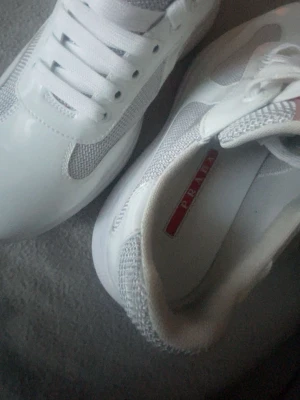 Vita sneakers från Prada med meshdetaljer - Snygga vita sneakers från Prada med klassisk rund tå och platt sula. Skorna har meshdetaljer på sidorna och ovandelen, samt snörning framtill. Insidan pryds av den ikoniska röda Prada-loggan. Materialmix av skinn och syntet för en clean look.