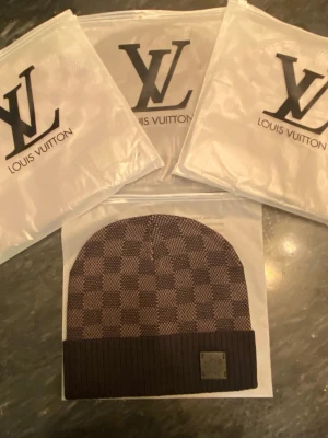 Svart Louis Vuitton Damier-mössa - Snygg Svart mössa från Louis Vuitton med klassiskt Damier-rutmönster och ribbad kant. Mössan har en metallbricka med Louis Vuitton-logga framtill. Perfekt accessoar för dig som vill ha en exklusiv och trendig look.