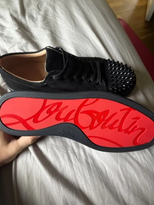  Svarta Christian Louboutin med spikar - Säljer ett par svarta sneakers från Christian Louboutin med ikonisk röd sula och coola svarta nitar på tån. Skorna har snörning och är tillverkade i mocka med läderdetaljer. Perfekta för dig som vill sticka ut med en edgy look.