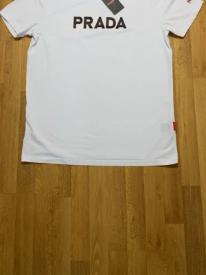 Vit  t-shirt med logga - Snygg vit t-shirt  med stor svart logga på bröstet och röd broderad  på ärmen. Klassisk passform och rund hals. Tillverkad i mjuk bomull för skön känsla. Röd detalj i nederkant ger extra premiumkänsla.