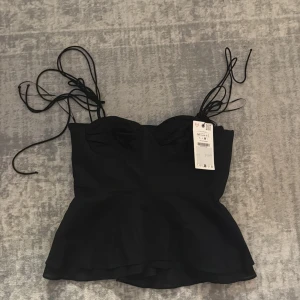 Svart topp med knytband - Trendig svart linnetopp från Zara med tunna knytband över axlarna och lätt utsvängd nederdel. Toppen har en söt bystdetalj och är perfekt för att skapa en chic look. Materialet känns mjukt och lätt, vilket gör den skön att bära. Helt ny med prislapp på!