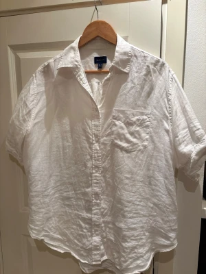Vit linneskjorta från Gant - Klassisk vit skjorta från Gant i svalt linne. Skjortan har korta ärmar, bröstficka och knappar framtill. Perfekt för en avslappnad och stilren look. Passar dig som gillar enkel och tidlös stil.