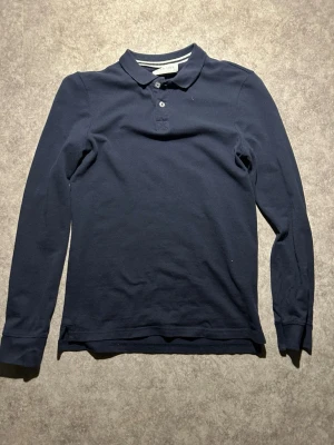 Marinblå långärmad pikétröja - Snygg marinblå långärmad pikétröja från Pier One med klassisk krage och knappar framtill. Tillverkad i mjuk bomull som känns skön mot huden. Perfekt för dig som gillar en stilren och enkel look.