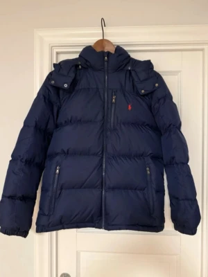  Ralph Lauren pufferjacka - Snygg marinblå pufferjacka från Polo Ralph Lauren med röd logga på bröstet. Jackan har dragkedja framtill, två sidofickor med dragkedja och hög krage med huva. Perfekt för kalla dagar och riktigt stilren look.