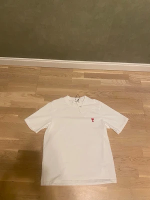 Vit t-shirt från Ami Paris med rött hjärta - Säljer en stilren vit t-shirt från Ami Paris med det ikoniska röda hjärta-A broderiet på bröstet. T-shirten har klassisk passform, rund halsringning och är tillverkad i mjuk bomull. Perfekt för en clean och trendig look.