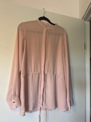 Ljusrosa blus  - Ljusrosa blus med lång ärm och knappar framtill. Blusen har ett tunt, mjukt tyg och ett knytband i midjan för en avslappnad men ändå feminin siluett. Storlek S från Gina Tricot. 