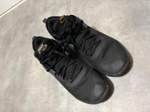 Svarta Nike sneakers med snörning - Säljer ett par helsvarta Nike sneakers med klassisk snörning och meshdetaljer för extra andningsförmåga. Skorna har en sportig och stilren look med Nike-logga i guld på plösen. Perfekta för dig som gillar en clean och enkel stil.