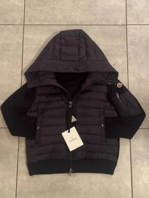 Moncler Cardigan marinblå - Säljer en svart pufferjacka från Moncler med stickade ärmar och rygg. Jackan har huva, dragkedja framtill och två fickor med tryckknappar. Klassisk Moncler-logga på ärmen och ribbade muddar för extra komfort. PRIS GÅR ATT DISKUTERAS