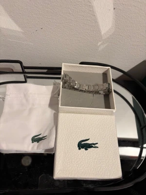 Silverfärgat armband från Lacoste - Snyggt armband i silverfärgad metall från Lacoste. Länkarna har ett rutigt mönster och den klassiska krokodilloggan syns på vissa länkar. Kommer i original ask och tygpåse med Lacoste-logga. Perfekt sophiad accessoar för dig som gillar stilrena detaljer.